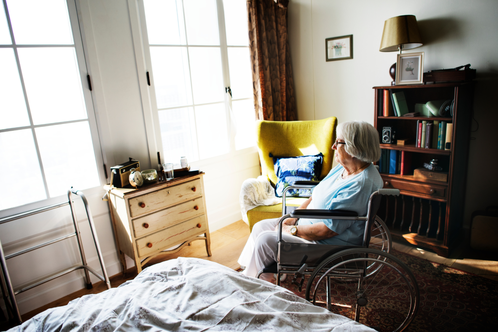 Image of an elderly white woman sitting in a wheelchair in a care facility room looking out the window